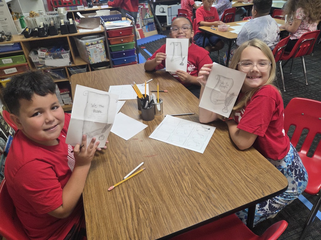 Three smiling elementary students sit around a classroom table, proudly holding up their illustrated drawings of the U.S. Constitution featuring the phrase "We the People." Pencils, activity sheets, and classroom storage shelves fill the surrounding space.