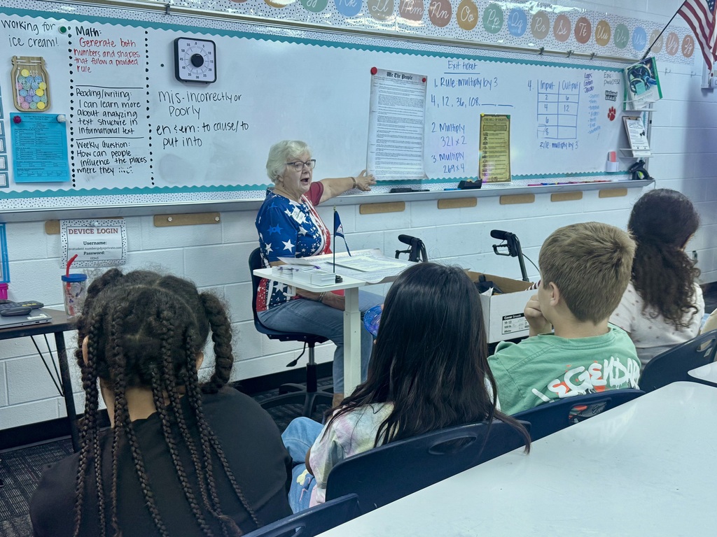 An adult guest speaker in patriotic apparel points toward a copy of the U.S. Constitution displayed on a whiteboard as elementary students watch attentively from their classroom desks. Colorful educational charts, learning objectives, and an alphabet strip line the board behind her.