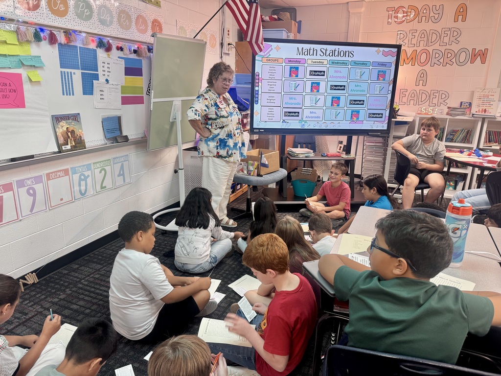 An adult guest speaker stands beside a large digital display showing a "Math Stations" schedule while addressing a class of elementary students gathered on the classroom carpet. Whiteboards with educational posters, place-value charts, and the slogan "Today a reader, tomorrow a leader" line the surrounding walls.