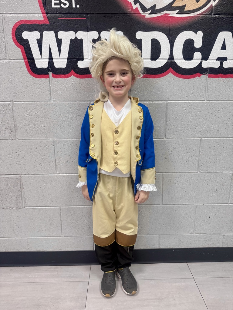 A young student dressed in a Colonial-era Founding Father costume—featuring a powder-blue jacket, tan vest and trousers, and a white powdered wig—smiles while standing in front of a school wall display.