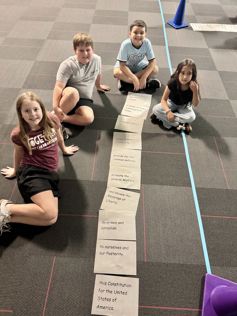 Four elementary students sit cross-legged on a carpeted floor, posing next to cards arranged in sequence containing lines from the Preamble to the U.S. Constitution.