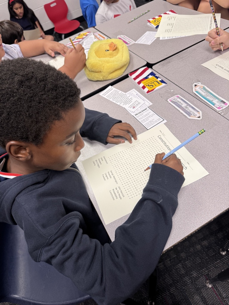 A young student sits at a classroom desk, holding a light blue pencil and working on a Constitution-themed word search activity sheet. Small educational bookmarks, pencil cases, and learning materials are organized on the surrounding desk surface.