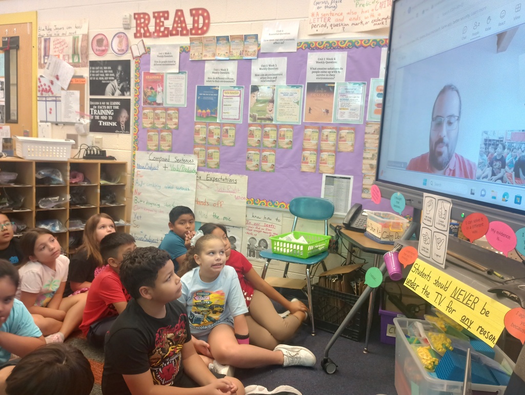 Main Subject: A group of elementary school students sitting on a blue carpet in a classroom, looking up toward a large digital display screen on the right. Screen Display: The screen shows a video call featuring a male presenter wearing a pink shirt and glasses. Classroom Environment: The background features educational bulletin boards with reading anchor charts, a large "READ" sign, cubbies with supplies, and a sign under the display reading, "Students should NEVER be under the T.V. for any reason."