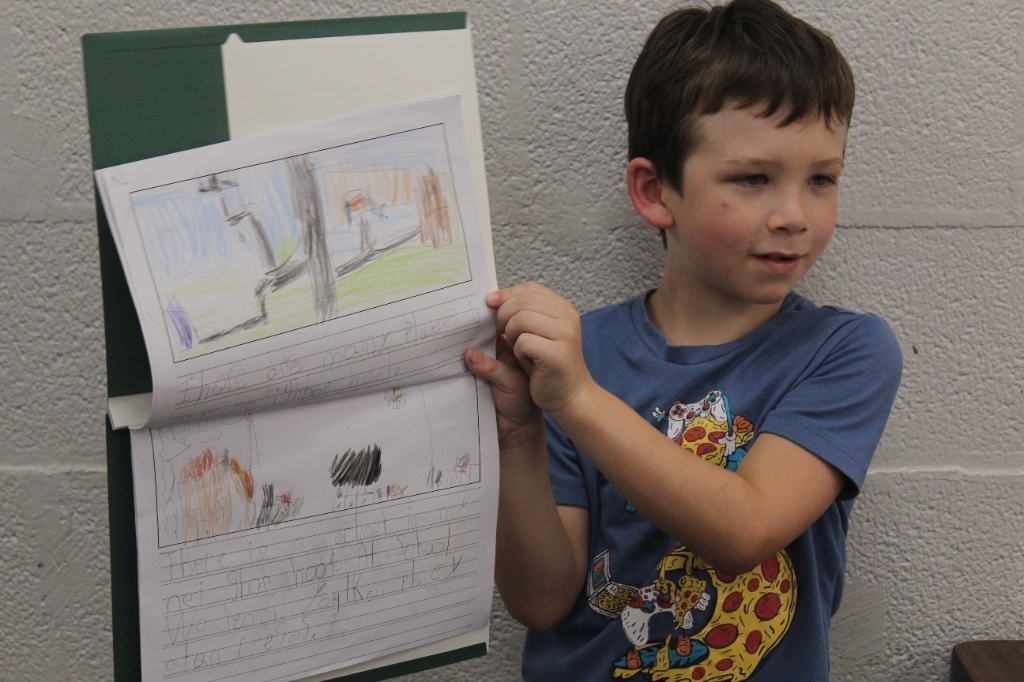 A young boy wearing a blue pizza-patterned t-shirt holding up a two-page handwritten story with colored drawings.