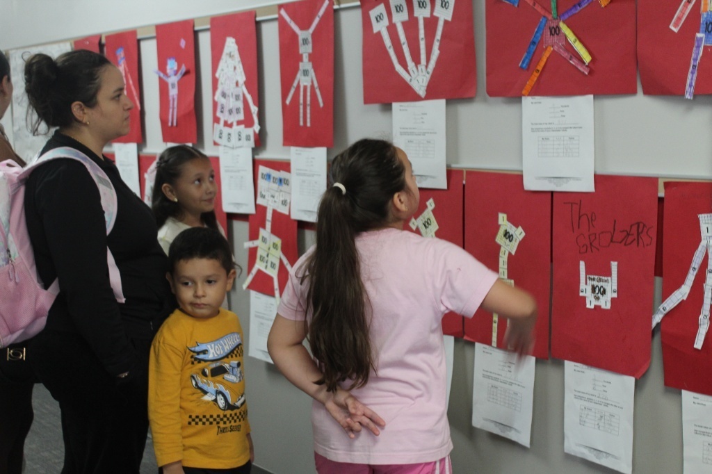 A mother and three children looking at colorful student math and artwork displays posted on a classroom wall.