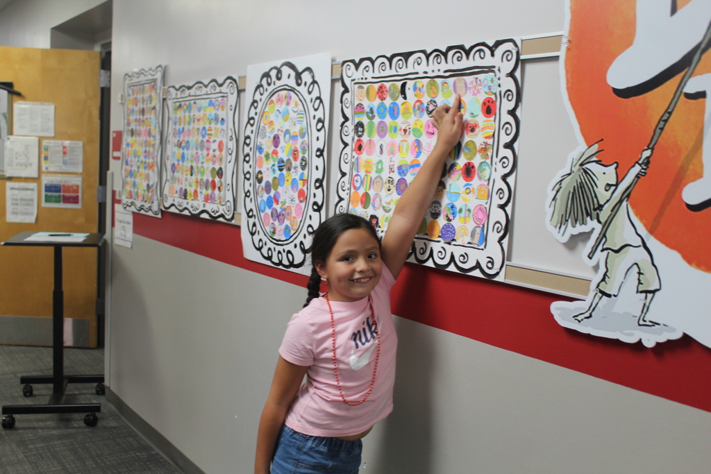 A young girl wearing a pink shirt and red necklace smiling as she points to artwork displayed on a hallway bulletin board.