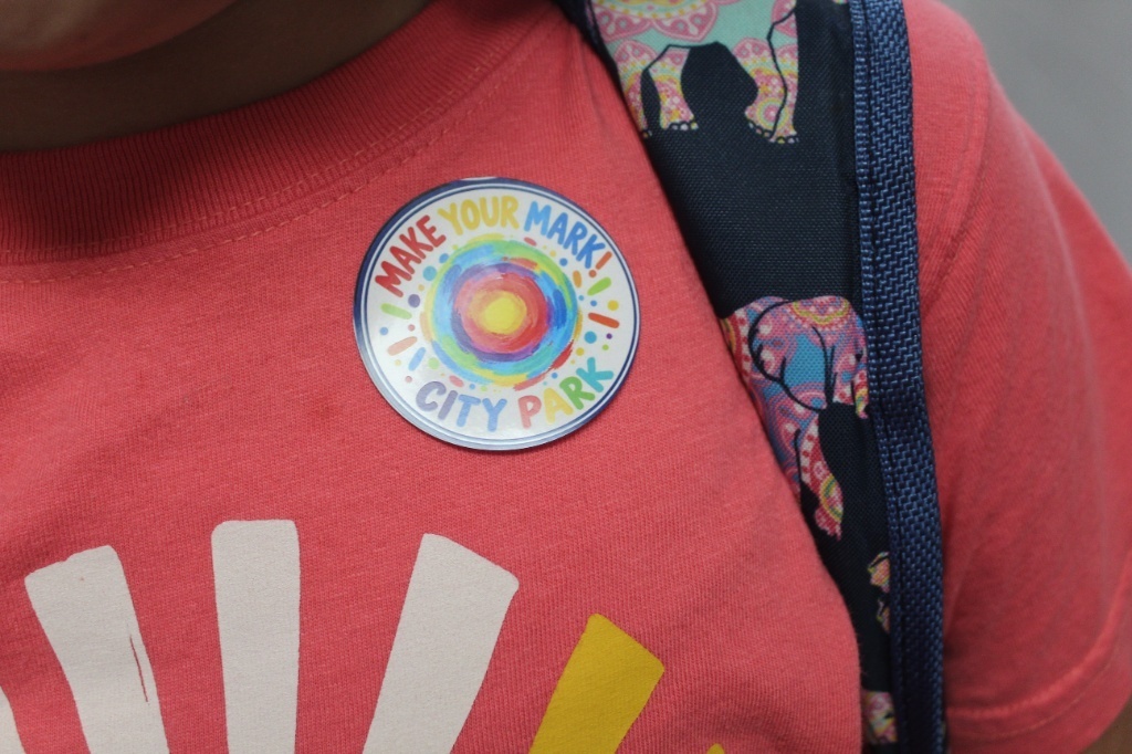 A close-up shot of a student wearing a circular sticker on their pink shirt that reads "MAKE YOUR MARK! CITY PARK" around a colorful, hand-drawn dot.