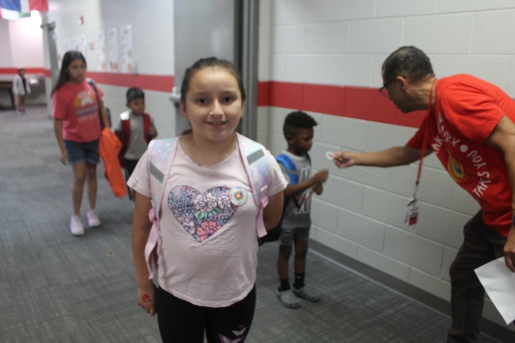 A young girl with a backpack smiling in a school hallway while wearing a "Make Your Mark!" sticker on her shirt. In the background, a staff member in a red shirt hands another student a sticker.