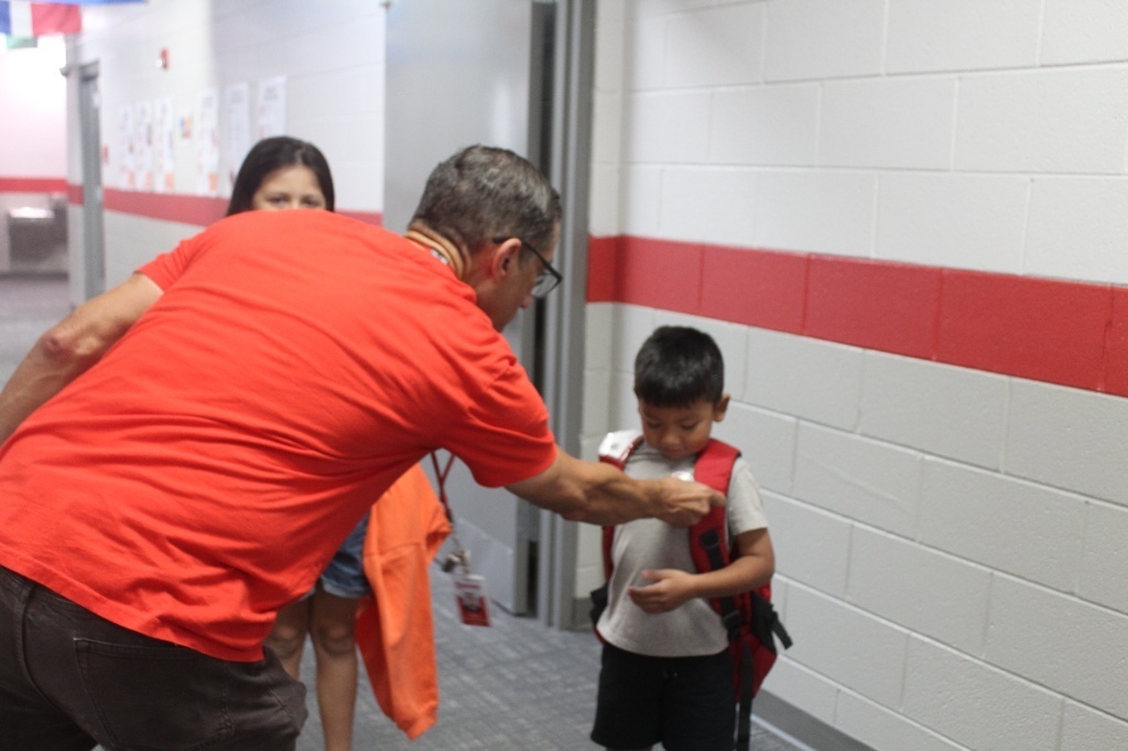 A staff member wearing a red shirt bends down to place a Dot Day sticker on a young student's backpack in a school hallway