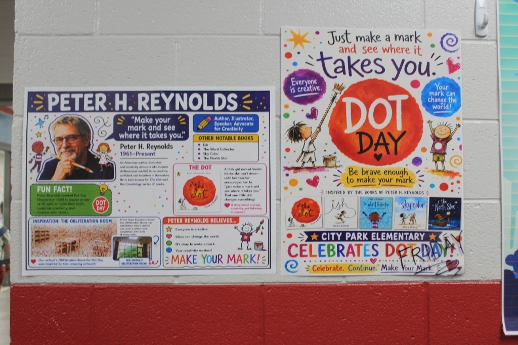 Two colorful posters on a white cinder block wall celebrating Dot Day and author Peter H. Reynolds. The poster on the left highlights facts about Reynolds, his books, and his belief to "Make Your Mark!". The poster on the right features artwork inspired by The Dot and reads "City Park Elementary Celebrates Dot Day."