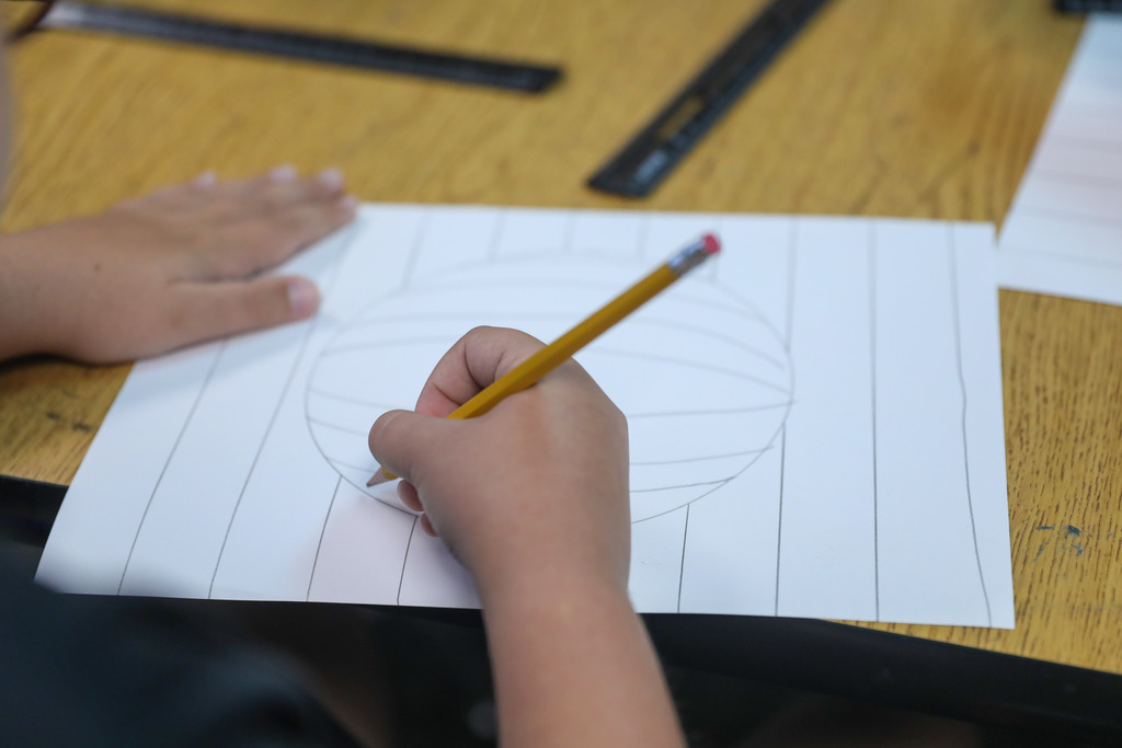 A close-up view of a student's hands drawing curved lines to create a 3D sphere effect on paper.