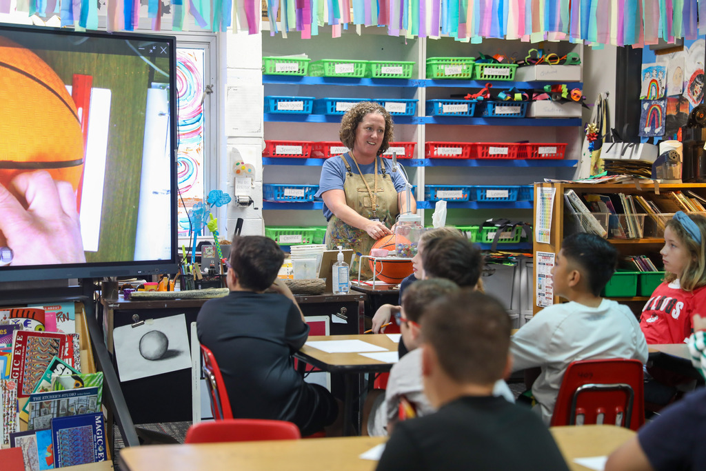An art teacher giving instructions to a class of elementary students watching a demonstration screen.