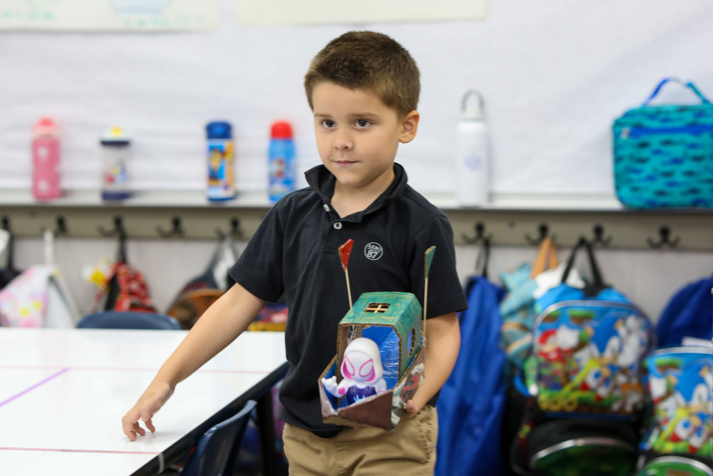 A student proudly holding up his handmade toy boat craft in a classroom.