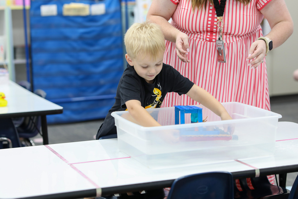 A student testing a craft-stick boat in a water tub under a teacher's guidance.
