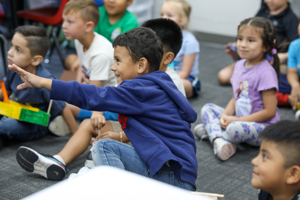 A young student raising his hand while sitting on the carpet with classmates.