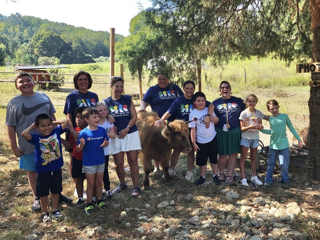 A large group of children and adults pose smiling outdoors next to a shaggy brown miniature cow in a sunny farm setting. Several adults in the group wear matching dark blue t-shirts featuring a colorful puzzle piece autism awareness design.