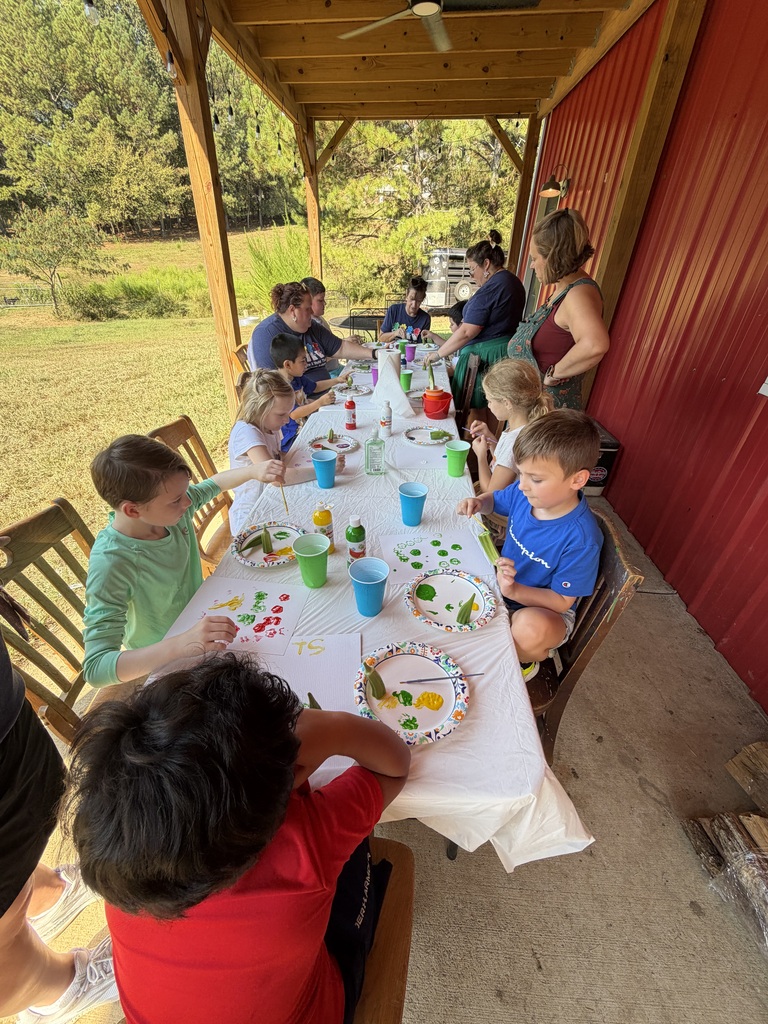 Children and adults sit at a long table covered with a white tablecloth under a covered wooden porch, participating in a outdoor painting activity with paper plates, paint cups, and brushes.