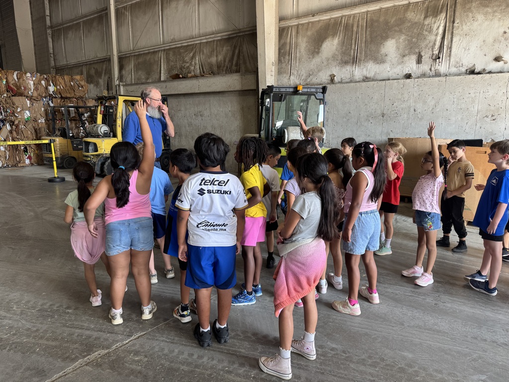 students in a group inside of the recycling facility listening to a worker share about his career while in front of a forklift. some students are raising their hands to answer questions