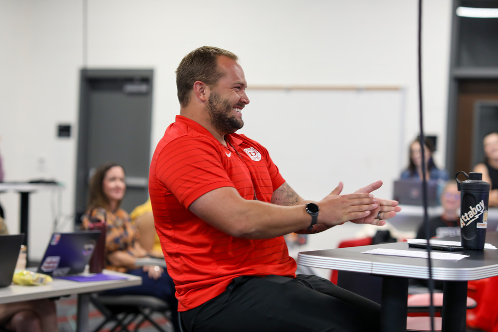 A male staff member sitting at a conference table laughing and clapping during a staff meeting.