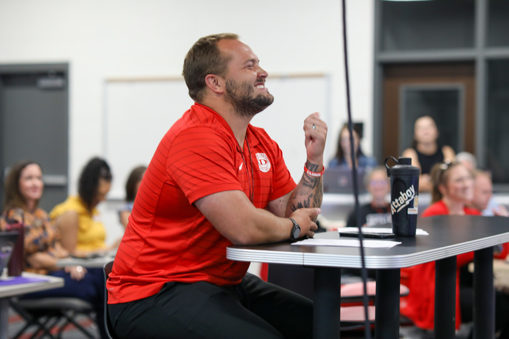 A male staff member sitting at a table smiling during a district presentation.