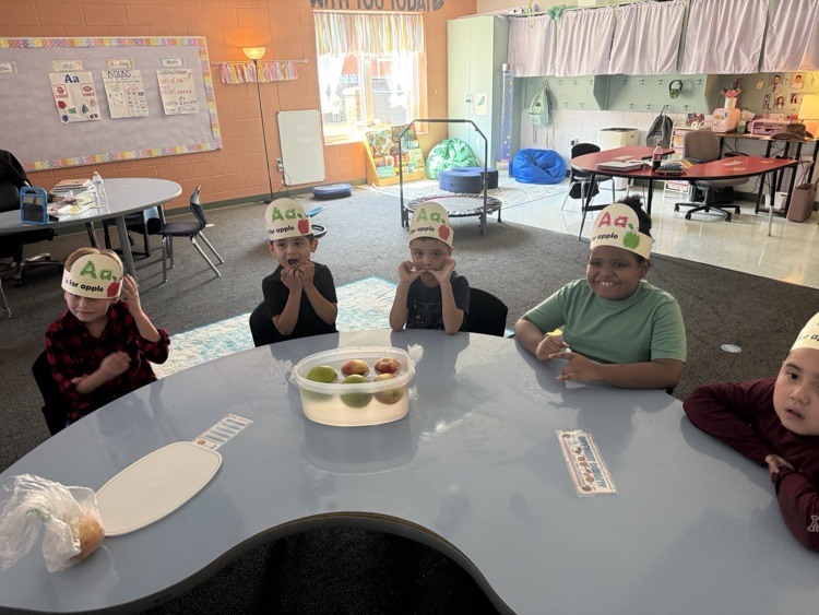Five young students sitting around a crescent-shaped table in a classroom, wearing paper crowns that say "Aa is for apple." They are participating in a lesson with a clear tub of floating apples in the center