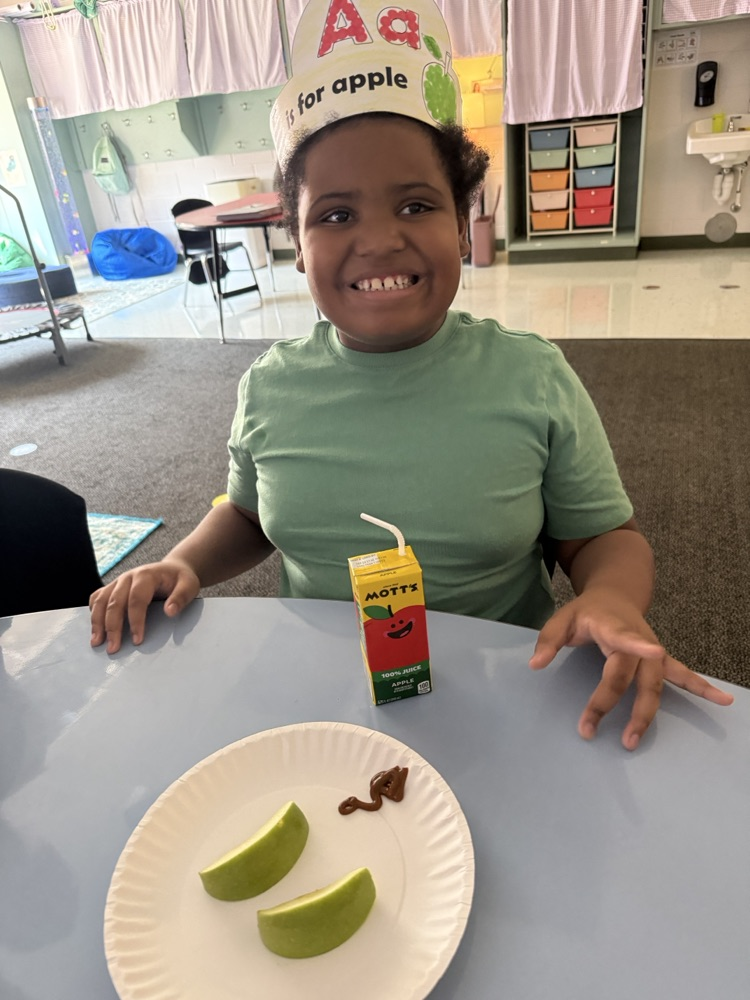 A young student smiling at a table while wearing an "Aa is for apple" paper headband, with a paper plate of green apple slices and a Mott's apple juice box in front of them