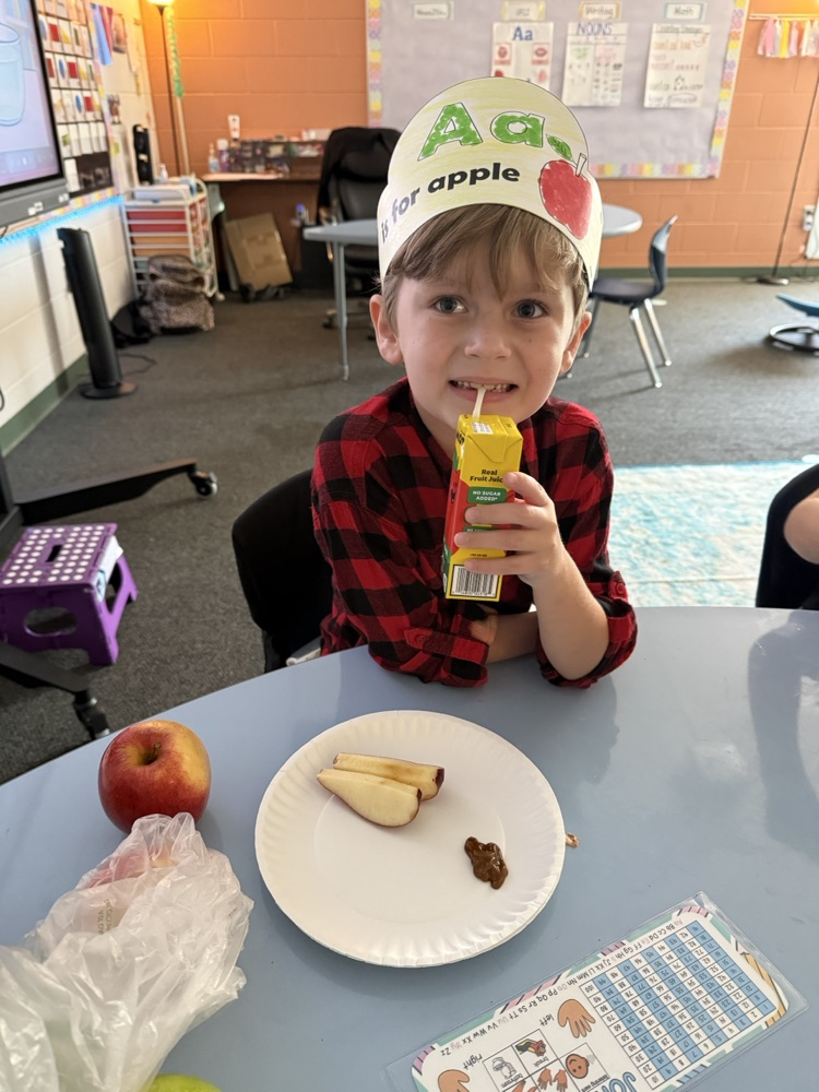 A young student smiling at a table while wearing an "Aa is for apple" paper headband, with a paper plate of green apple slices and a Mott's apple juice box in front of them