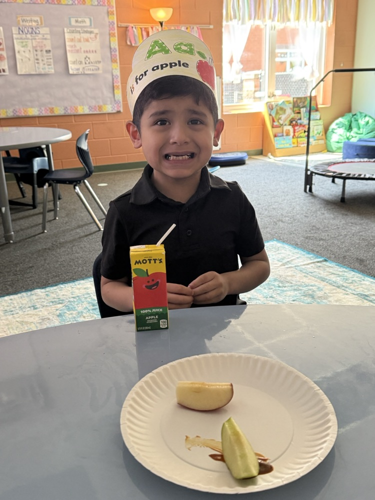 A young student smiling at a table while wearing an "Aa is for apple" paper headband, with a paper plate of green apple slices and a Mott's apple juice box in front of them
