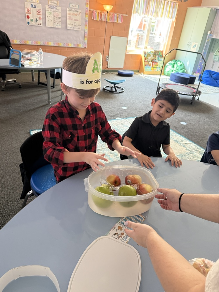 A close-up of a young boy wearing a red plaid shirt and an "Aa is for apple" paper headband, drinking from a Mott's apple juice box next to a plate of sliced apples