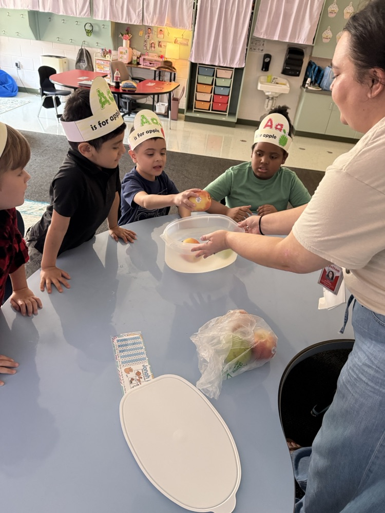 A teacher assisting young students as they place apples into a container of water during a classroom learning activity. All students are wearing "Aa is for apple" paper headbands
