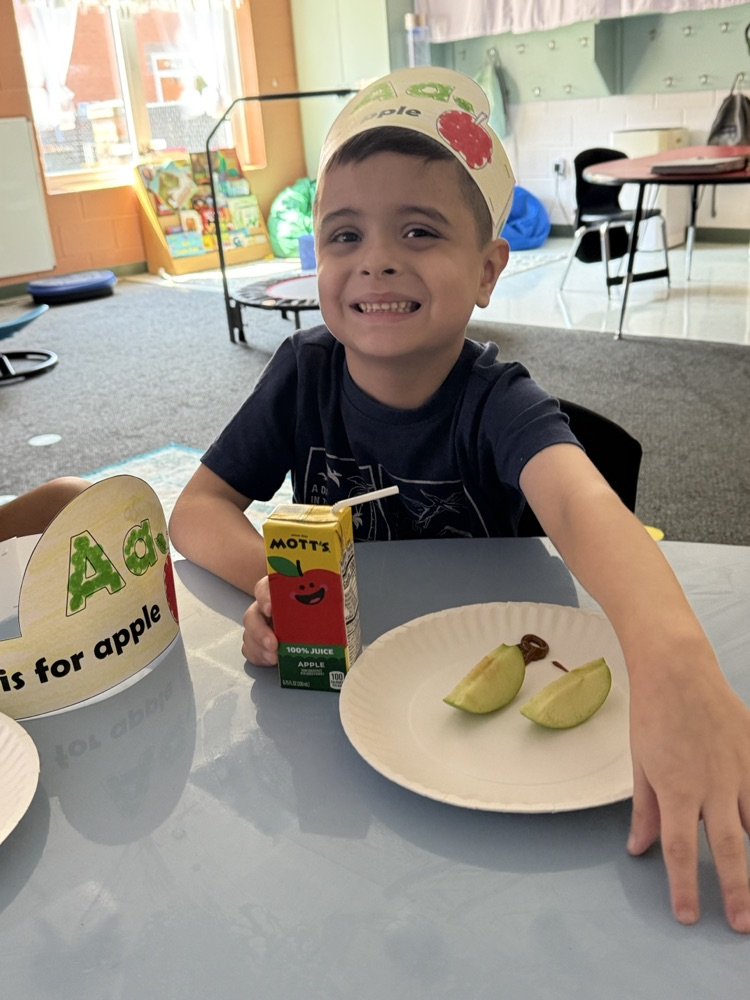 A young boy wearing an "Aa is for apple" headband smiling at the camera, sitting at a table with a plate of apple slices and an apple juice box