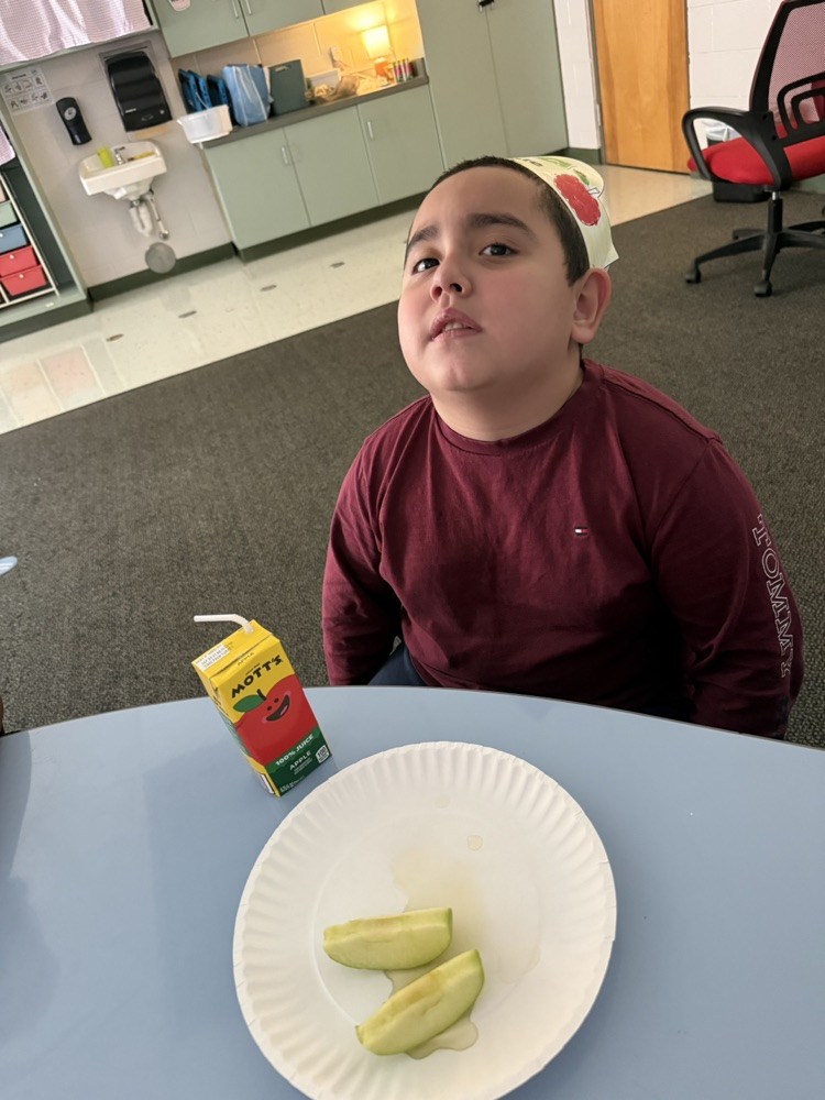 A student in a maroon long-sleeve shirt and paper headband sitting at a table with green apple slices on a paper plate and a Mott's apple juice box