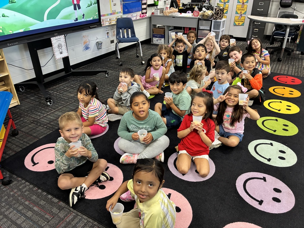 A group of young elementary students sitting cross-legged on a classroom rug patterned with colorful smiley face circles. The children are holding small plastic cups with snacks and smiling towards the camera, with a large interactive screen and classroom furniture in the background.