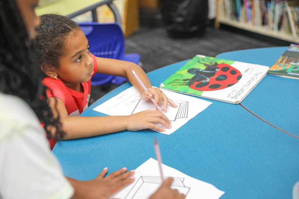 Young elementary students working on drawing and writing activities at a library table.