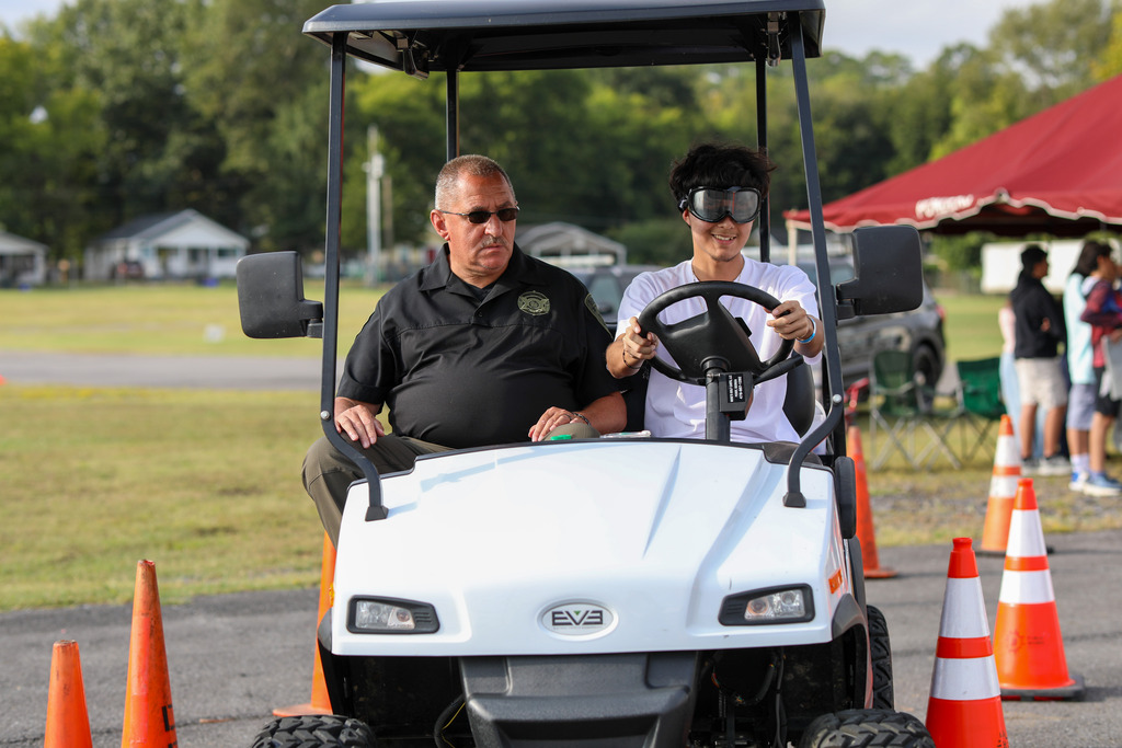 A student wearing impaired-vision goggles driving a utility cart alongside a safety officer.