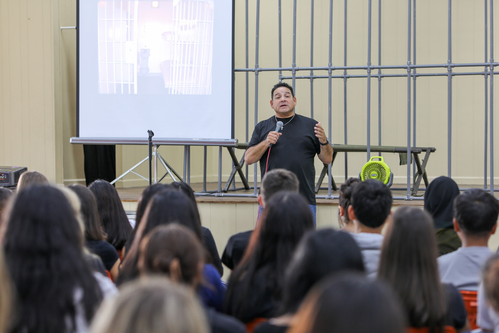 A guest speaker holding a microphone while addressing a large audience of seated students.