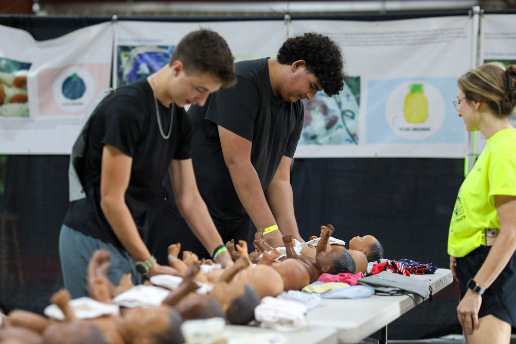 High school students learning infant care techniques using realistic doll models at an instructional booth.