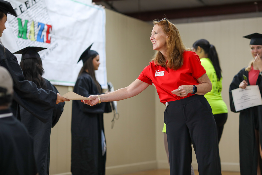 A school staff member handing out mock diplomas to students dressed in graduation caps and gowns during a Teen Maze activity.