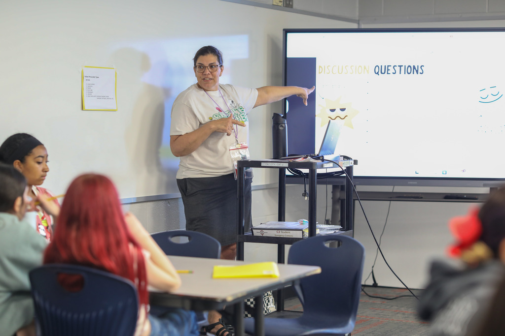 A teacher gesturing toward a screen displaying discussion questions for the class.