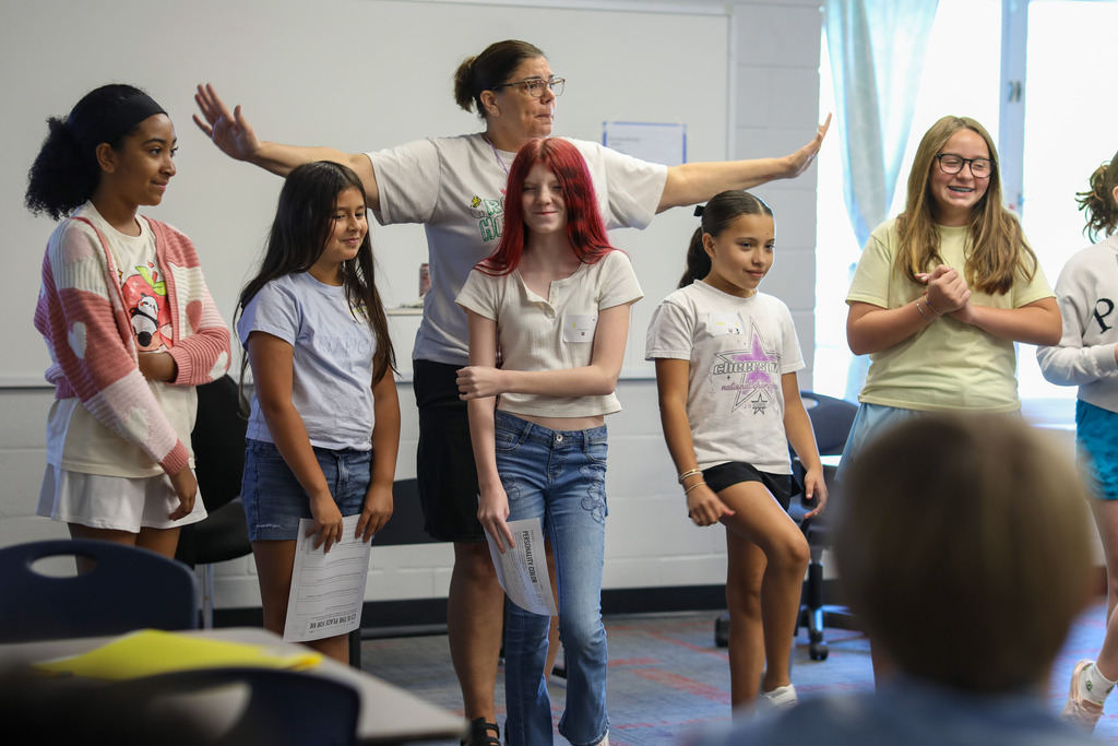 A teacher with outstretched arms leading a group presentation activity with students.