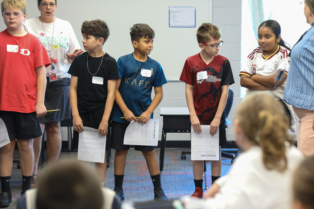 Students lined up along a classroom wall holding assignment papers.