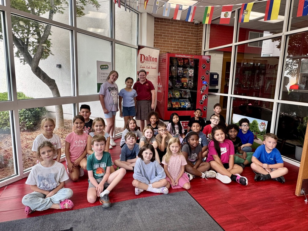 A large group of elementary school students posing and smiling together inside a school entryway with floor-to-ceiling windows, red flooring, international flags hanging above, and a book vending machine in the background.