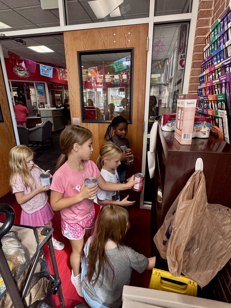 A group of young elementary students placing donated canned goods and non-perishable food items onto shelves near a resource center shelf inside a school entryway.