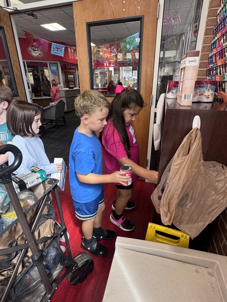 Three students smiling standing behind a brown folding wagon filled with donated canned food, non-perishable groceries, and hygiene supplies inside a school hallway.
