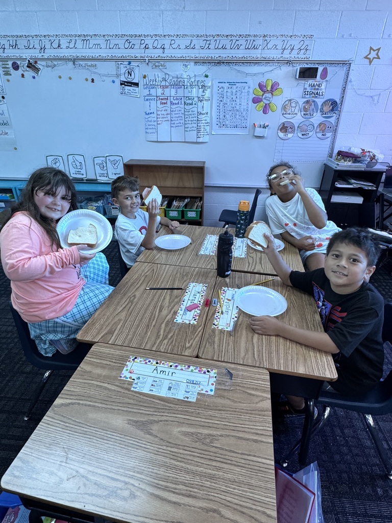Four young students gathered around a cluster of classroom desks smile at the camera while holding up paper plates and sandwiches during a classroom activity.
