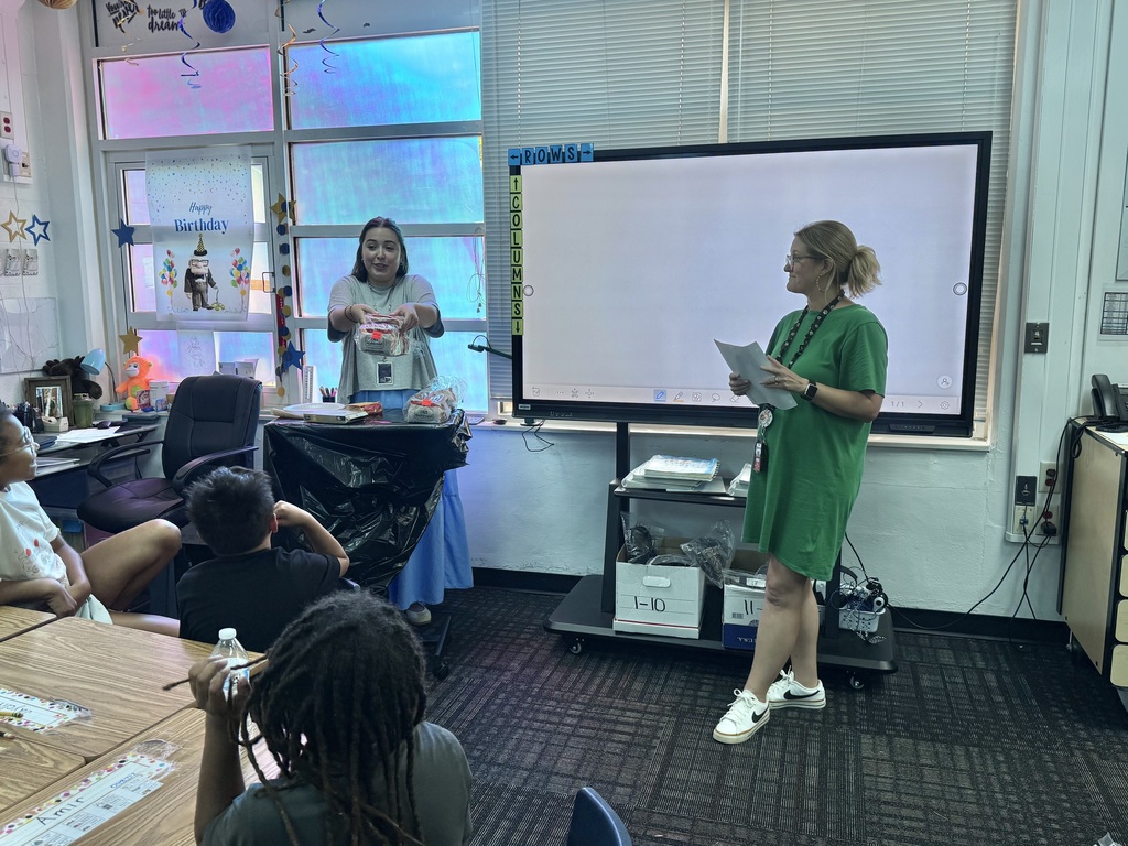 Two teachers lead a presentation at the front of a classroom next to an interactive display screen. One teacher in a green dress holds papers while the other hands out items to students seated at desks in the foreground.