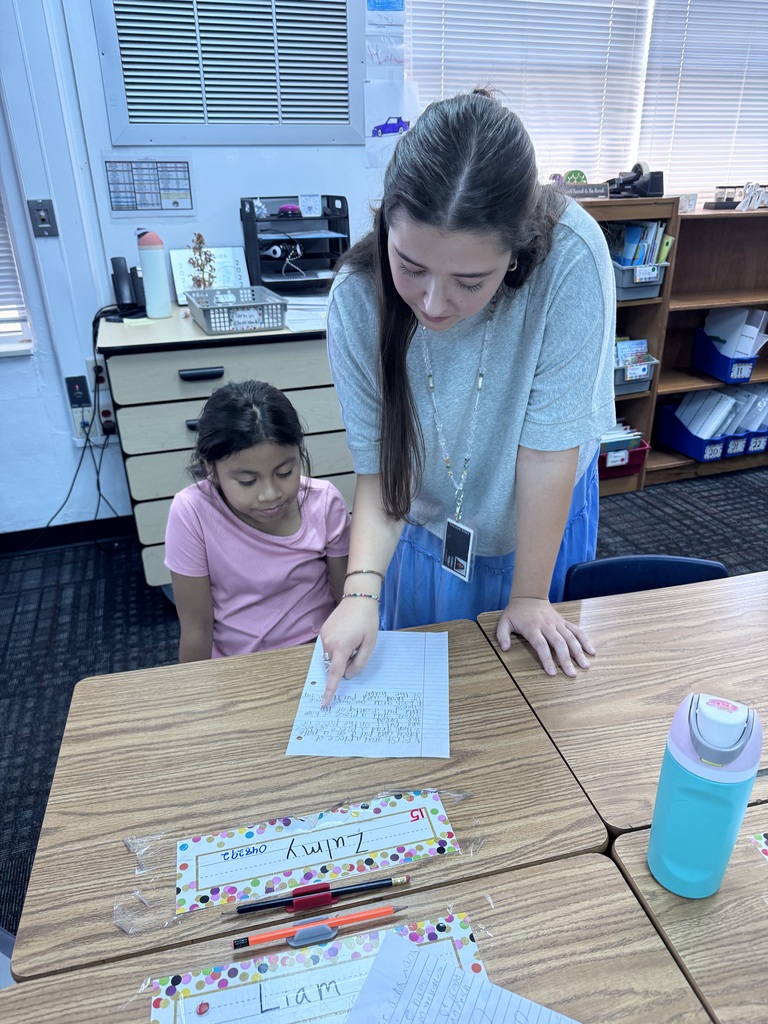 A female teacher wearing a light blue top leans over a desk to point at a student's handwritten assignment, helping a young female student seated beside her.