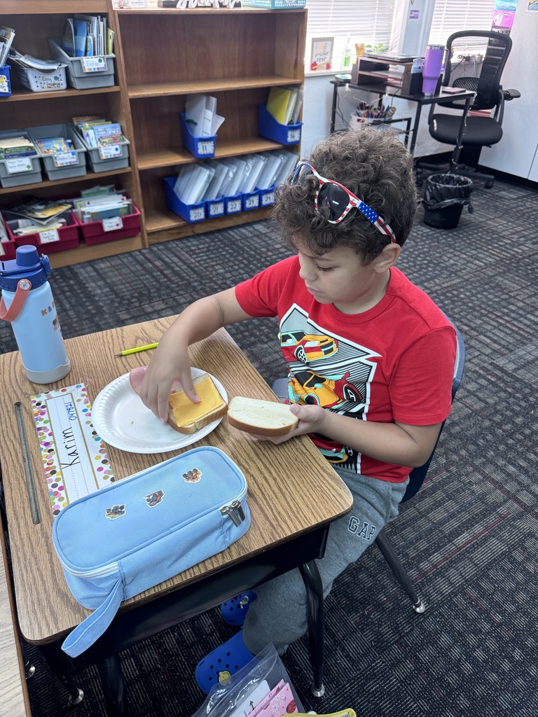 A young boy in a red graphic t-shirt and sunglasses pushed up on his head sits at his classroom desk assembling a ham and cheese sandwich on a paper plate.
