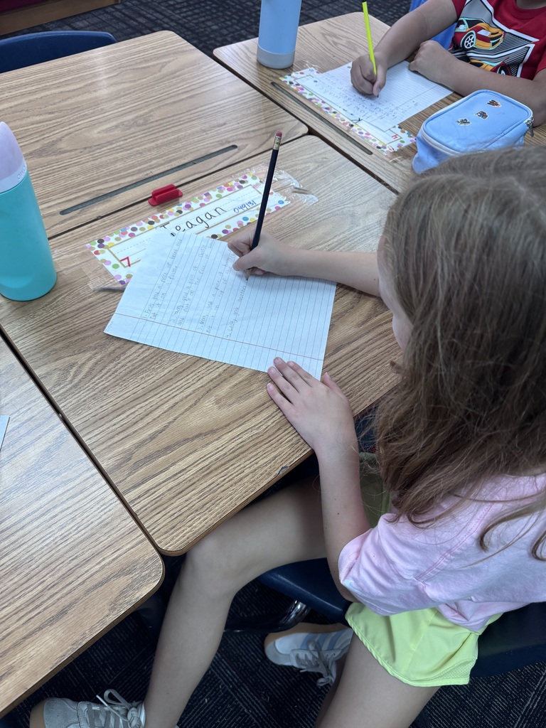 An over-the-shoulder view of a young student in a pink shirt and light yellow shorts writing on lined paper with a pencil at a wooden classroom desk.