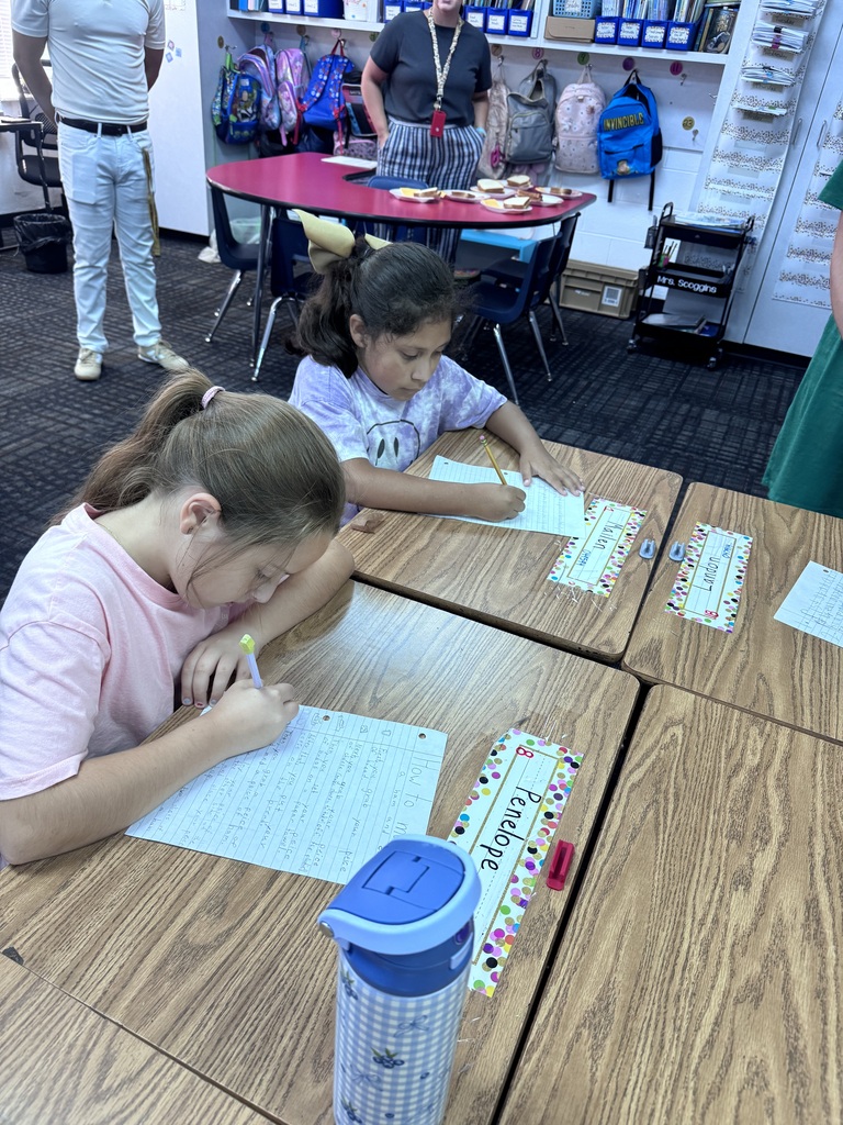 Two young girls sit at adjoining wooden desks in a classroom, focused on writing step-by-step instructions on lined paper while adults stand nearby in the background.